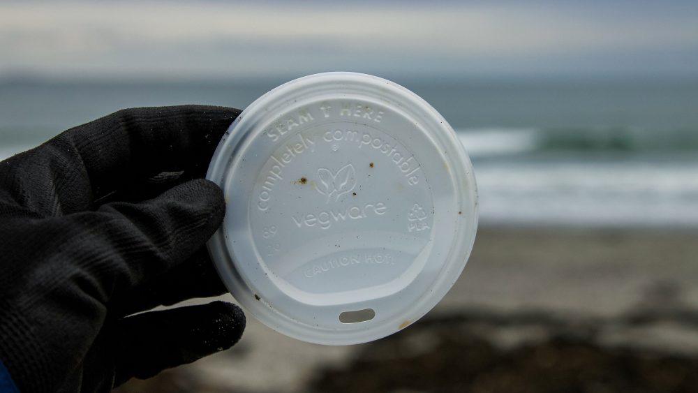 A hand holding a compostable plastic lid with a beach in the background