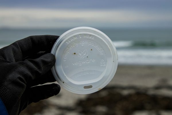 A hand holding a compostable plastic lid with a beach in the background